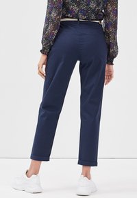 Femme portant un pantalon bleu marine retroussé, des baskets blanches et un haut à manches longues à motif floral, debout devant un fond blanc uni.