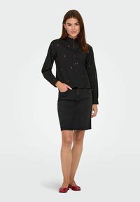 Donna in blusa nera con bottoni e piccoli motivi floreali rossi, gonna di jeans nera e scarpe rosse basse, posa con una mano in tasca.
