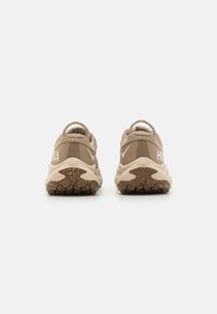 Beige athletic shoes with a textured upper, reflective accents, and a rugged brown outsole. Features pull tabs for easy wear.