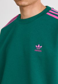 Green sweatshirt with pink accent stripes on the shoulders and a small pink Adidas logo on the chest. Soft cotton material, relaxed fit.
