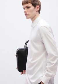 Μαύρη δερμάτινη τσάντα με φερμουάρ, κρατημένη στο χέρι. Το άτομο φοράει εφαρμοστό λευκό πουκάμισο με κουμπιά και μακριά μανίκια.