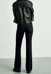 Jeans de mezclilla oscuros acampanados con cintura alta y sutiles detalles de costura, combinados con una chaqueta de cuero negra corta. Posicionados desde atrás.