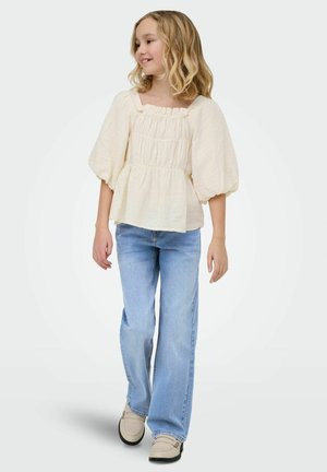 Fată tânără zâmbitoare, purtând o bluză crem cu mâneci bufante, blugi albastru deschis și pantofi bej, stând în fața unui fundal simplu.