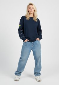 Una donna bionda sta in piedi con le mani in tasca, indossa un felpa blu navy con maniche ricamate, jeans larghi di un blu chiaro e sneakers bianche.