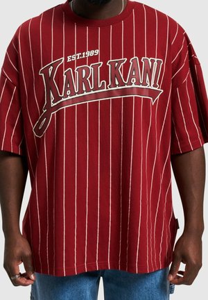 Muž nosící volné červené tričko s tenkými bílými svislými proužky a nápisem "KARL KANI EST. 1989" přes hrudník.