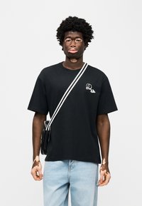 Μαύρο βαμβακερό t-shirt με στρογγυλή λαιμόκοψη, κοντά μανίκια και λευκό κεντητό λογότυπο στο στήθος, σε συνδυασμό με ανοιχτόχρωμο μπλε τζιν και μαύρη τσάντα.