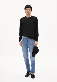 Schwarzes Langarm-Sweatshirt, hellblaue Jeans, schwarze Mütze in der Hand und schwarze Schuhe. Schlichtes Design mit lässiger Passform und glatter Textur.