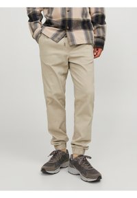 Beiget puuvillaiset jogger-housut, joissa on elastiset resorit ja kiristysnyöri vyötäröllä, yhdistetty harmaisiin lenkkareihin. Kangas näyttää sileältä ja vähäpintaiselta.