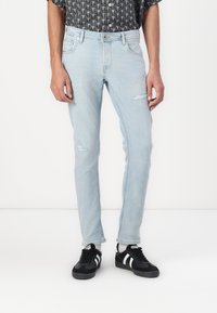 Svijetloplavi traperice slim fit kroja, s istrošenim detaljima i srednjim strukom, uparen s crnim tenisicama i košuljom s uzorkom.
