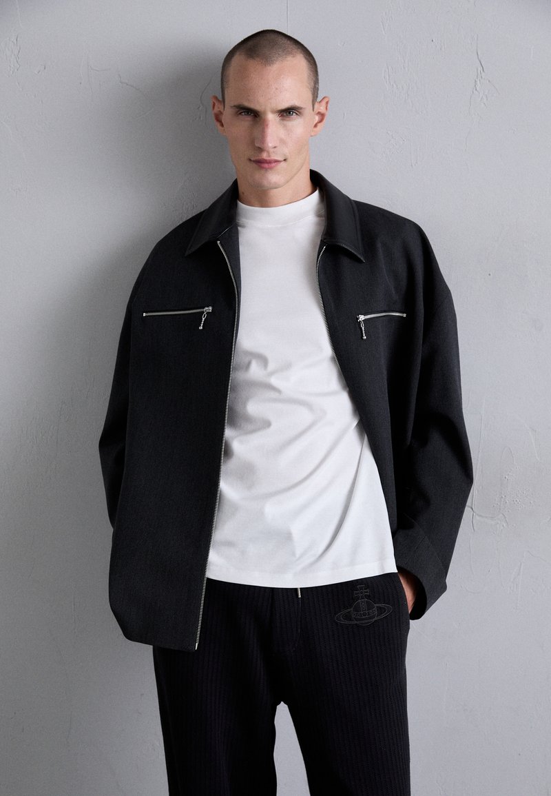 Veste zippée noire avec deux poches avant, portée sur un t-shirt blanc. Pantalons rayés sombres avec un design de logo subtil près de la poche.