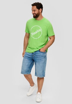 Grünes Baumwoll-T-Shirt mit einem weißen, runden "Sinalco"-Logo, kombiniert mit hellblauen Denim-Shorts und weißen Sneakern. Lässiges Design.