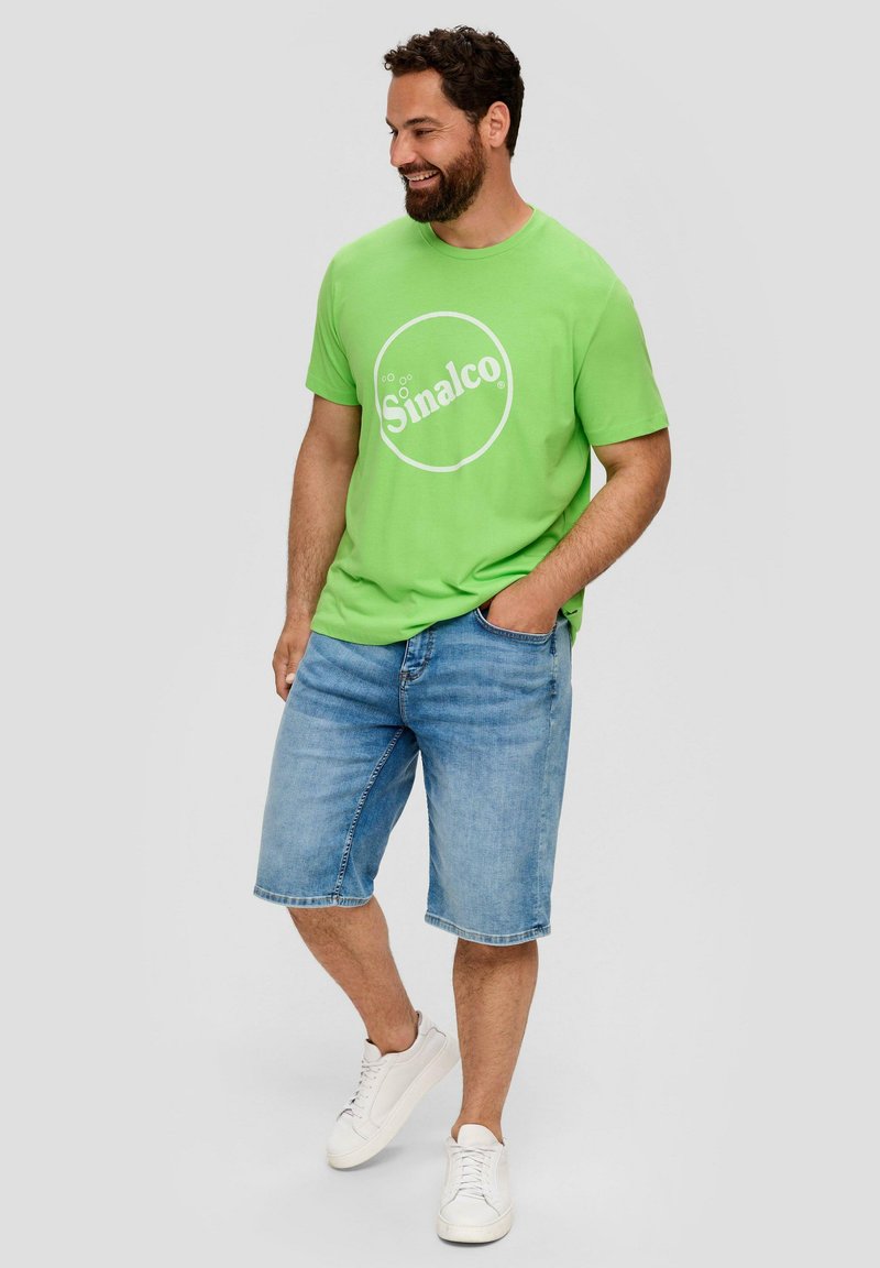 Groene katoen t-shirt met een wit cirkelvormig "Sinalco" logo, gecombineerd met lichtblauwe denim shorts en witte sneakers. Casual ontwerp.