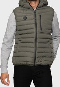 Veste sans manches matelassée en vert olive, avec une fermeture éclair frontale et une poche poitrine. Caractérisée par une surface texturée et une capuche, portée avec un pull gris.