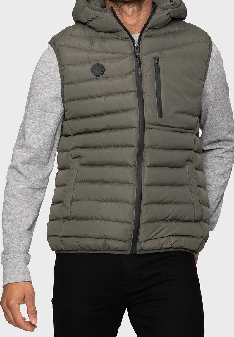 Veste sans manches matelassée en vert olive, avec une fermeture éclair frontale et une poche poitrine. Caractérisée par une surface texturée et une capuche, portée avec un pull gris.