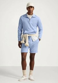 Άνδρας που φοράει ανοιχτό μπλε φούτερ με κοντό φερμουάρ και ταιριαστά σορτς, κρεμ πουλόβερ που είναι δεμένο στη μέση, κρεμ καπέλο, κάλτσες και λευκά αθλητικά παπούτσια.