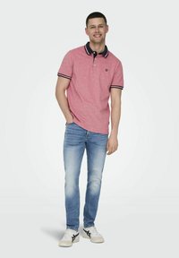 Rode polo met een marineblauwe kraag en gestreepte afwerking, gecombineerd met lichtblauwe jeans en witte sneakers. Het model staat met een ontspannen uitstraling.