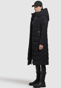Sort pufferjakke med lang design, chevron-quiltet mønster, høj krave, sidelomme med lynlås og et mærketag. Matches med sorte leggings og støvler.