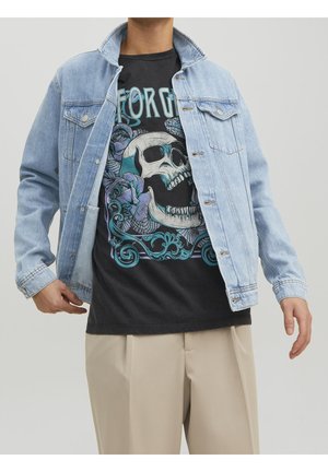 Osoba nosząca jasnoniebieską kurtkę jeansową na czarnym t-shircie z dużym nadrukiem czaszki i niebieskich kwiatów, w połączeniu z beżowymi spodniami.