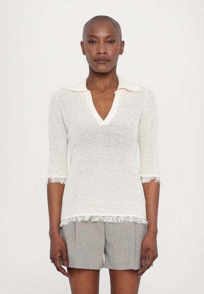 Knit top in off-white with a V-neck, short sleeves, and fringe accents at the cuffs and hem. Paired with gray tailored shorts.