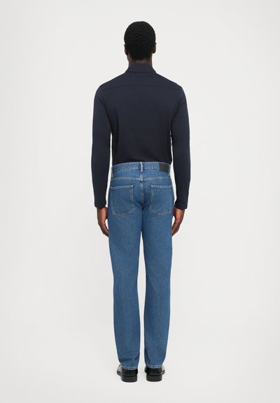 Man standing facing away wearing a navy long-sleeve shirt, blue straight-leg jeans, and black shoes against a plain light background.