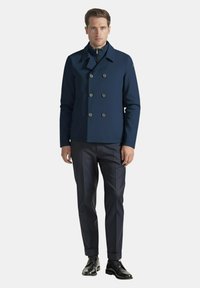 Uomo che indossa un giacca doppiopetto blu navy, pantaloni sartoriali scuri e scarpe eleganti nere lucidate, in piedi contro uno sfondo neutro.