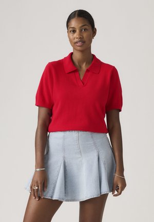 Nő piros rövid ujjú galléros felsőt és világoskék redőzött farmer szoknyát visel, egyszerű háttér előtt áll.