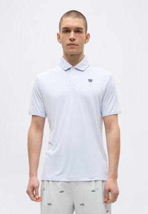 Giovane uomo che indossa una polo bianca semplice a maniche corte con un piccolo logo sul petto, in piedi davanti a uno sfondo uniforme.