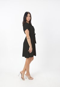 Donna con lunghi capelli scuri, vestita con un abito nero a bottoni e scarpe con tacco trasparenti, in piedi di profilo su uno sfondo bianco.