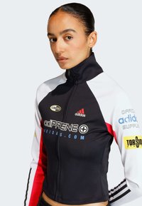Crna, bijela i crvena atletska jakna s visokim ovratnikom. Sadrži logotipe, prednji zip i prilagođeni dizajn. Izrađena je od glatkog, rastezljivog materijala.