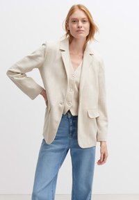Blazer beige con vestibilità comoda, due tasche frontali, chiusura con un solo bottone e tessuto testurizzato, abbinato a un gilet leggero e jeans blu.
