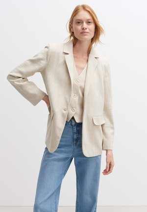 Beige blazer with a relaxed fit, two front pockets, single-button closure, and textured fabric, paired with a light vest and blue jeans.