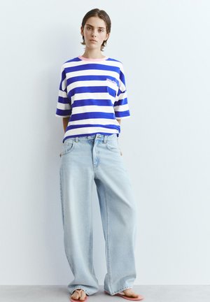 Mladá osoba stojaca pri bielej stene, oblečená v modro-bielo pruhovanej košeli a svetlomodrých dlhých džínsach, ruky v vreckách.