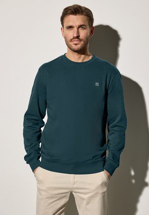 Dunkelblaues Sweatshirt mit langen Ärmeln, runden Halsausschnitt, weichem Stoff und kleinem Patch-Logo auf der Brust, kombiniert mit hellen Hosen.