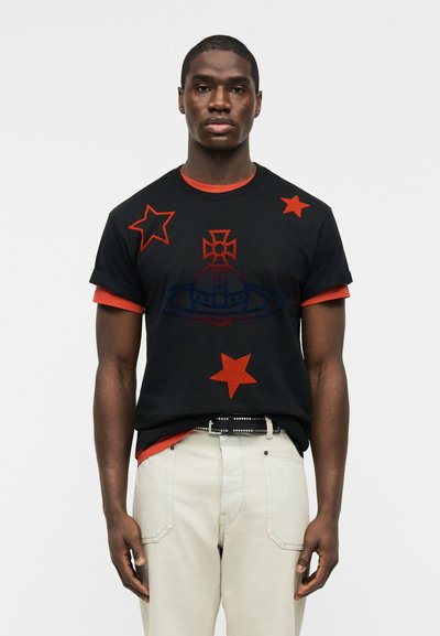 Άνδρας που φοράει μαύρο μπλουζάκι με κόκκινα αστέρια και έμβλημα, πάνω από κόκκινο πουκάμισο, συνδυασμένο με ανοιχτό μπεζ παντελόνι και μαύρη ζώνη με καρφιά.