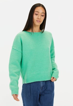 Mintgroene, gebreide trui met een losse pasvorm, geribbelde manchetten en onderrand, met een ronde halslijn en gevallen schouders. Draagt bij donkere blauwe broek.