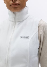 Κοντινό πλάνο ατόμου που φοράει λευκό γιλέκο Columbia από φλις, με το φερμουάρ κλειστό πάνω από μια μπεζ μακρυμάνικη μπλούζα με υφή.