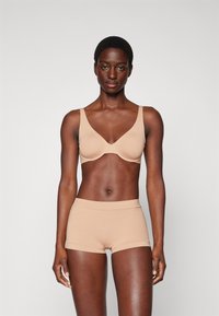 Nude-farvet bh med glatte skåle og brede stropper, parret med matchende sømløse boyshorts. Begge items har en blød tekstur og minimalistisk design.