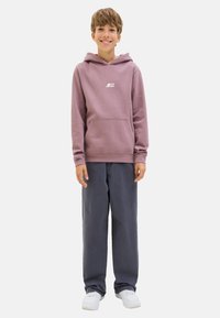 Un garçon souriant se tient dans un sweat à capuche violet et un pantalon gris, portant des baskets blanches, devant un fond blanc uni.