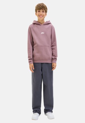 Glunderende jongen staat in een paarse hoodie en grijze broek, met witte sneakers, tegen een eenvoudige witte achtergrond.
