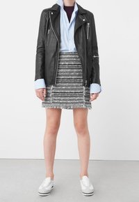 Veste de biker en cuir noir, chemise bleu clair, jupe texturée en tweed noir et blanc avec des franges, et baskets grises. Tenue simple et superposée.