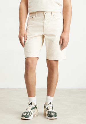 Shorts en denim couleur crème, coupe droite, cinq poches, ourlets retroussés, associés à des baskets vertes et blanches et des chaussettes blanches.