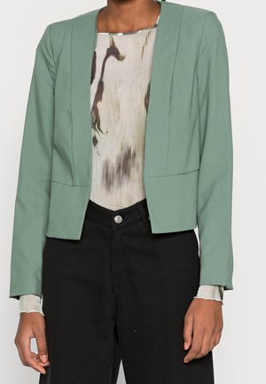 Personne portant un blazer ouvert vert clair sur un haut beige à motifs abstraits et un pantalon noir taille haute, debout devant un fond uni.