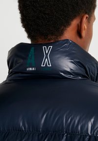 Doudoune marine à col haut. Présente les logos en relief "AX" et "Armani Exchange" dans le dos. Fini lisse et brillant, texture matelassée.