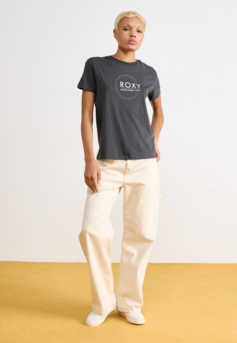 Camiseta de algodón gris con el logo blanco "ROXY", combinada con pantalones holgados crema y zapatillas blancas sin cordones, de pie contra un fondo claro.