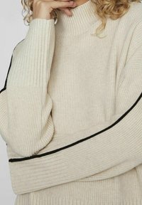 Beige geribbelde coltrui met zwarte accenten langs de mouwen, gemaakt van een dikke, gestructureerde breisel en een losse pasvorm.