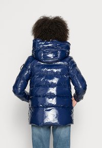 Person with curly hair wearing a shiny navy blue puffer jacket with hood, standing with hands on hips, facing away.