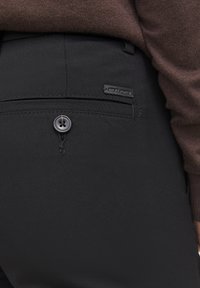Poche arrière d’un pantalon noir avec un bouton et une petite étiquette Jack & Jones, porté avec un pull marron.
