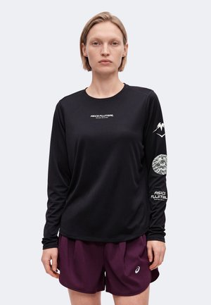 Nainen seisoo kasvot eteenpäin, päällään musta pitkähihaisen ASICS FUJITRAIL-paita ja violetit urheilushortsit yksinkertaisella vaalealla taustalla.