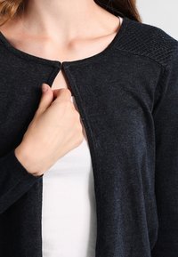 Cárdigan azul marino oscuro, material de punto suave, cuello redondo, cierre único en la parte delantera, acentos acanalados en los hombros, textura ligera.