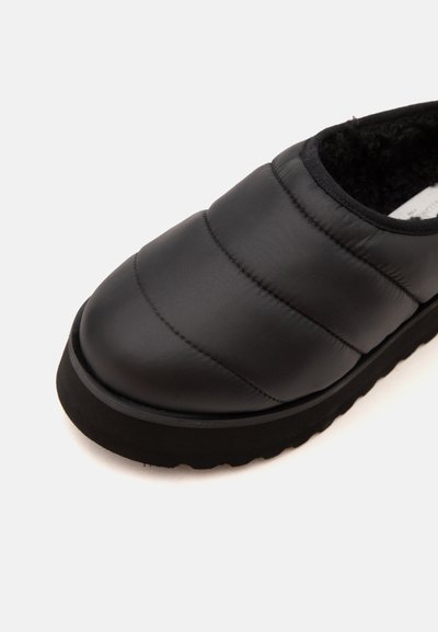 Μαύρο παπούτσι τύπου slip-on με παχιά σόλα και μαλακή εσωτερική επένδυση, εμφανίζεται από κοντινή γωνιακή άποψη σε λευκό φόντο.